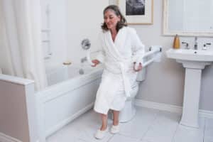 Photo of a woman wearing a white bathrobe using the upper length of two HealthCraft PT Rails to lower herself onto a closed toilet