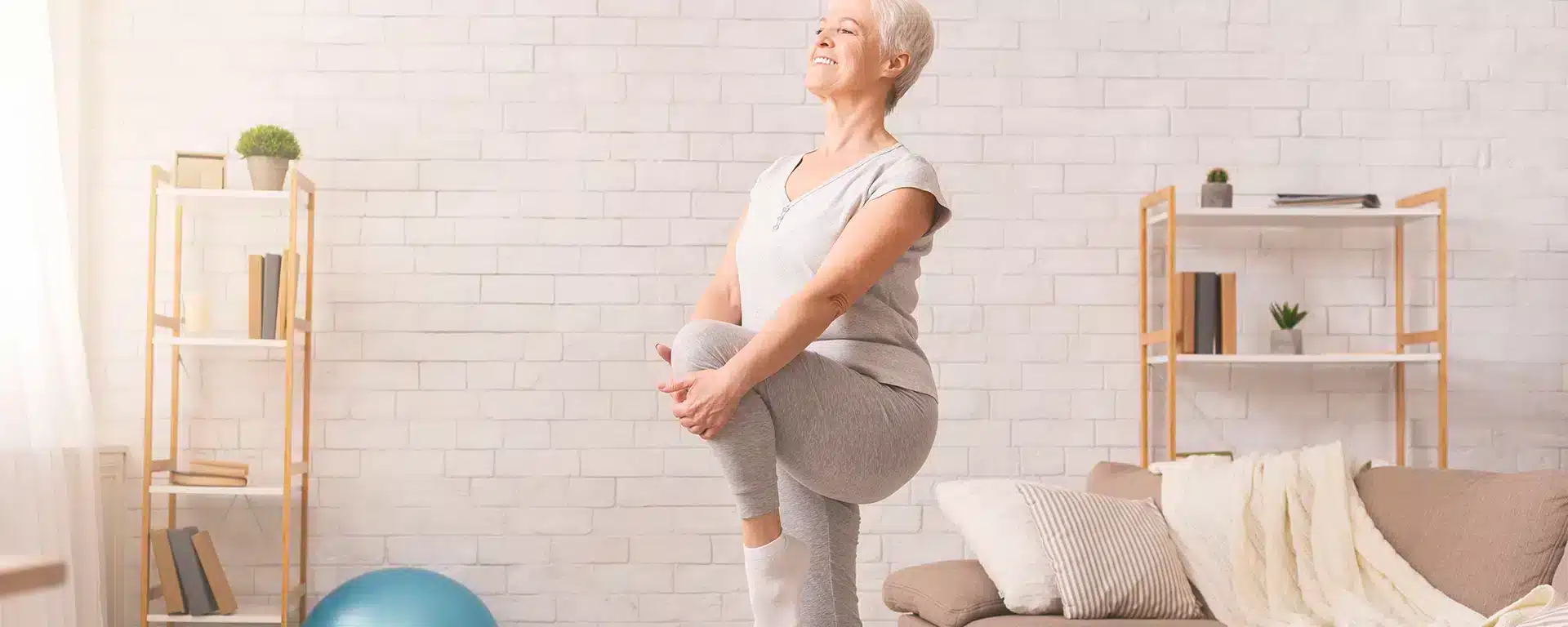 An elderly woman standing & stretching on one leg by pulling the other leg upwards at the knee.